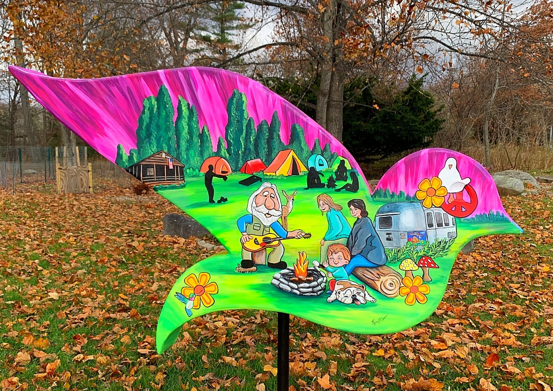 A whimsical, bird-shaped wooden sign painted with a vibrant camping scene featuring a family around a campfire, colorful tents, a vintage trailer, and a pink sky, set against a backdrop of autumn leaves.