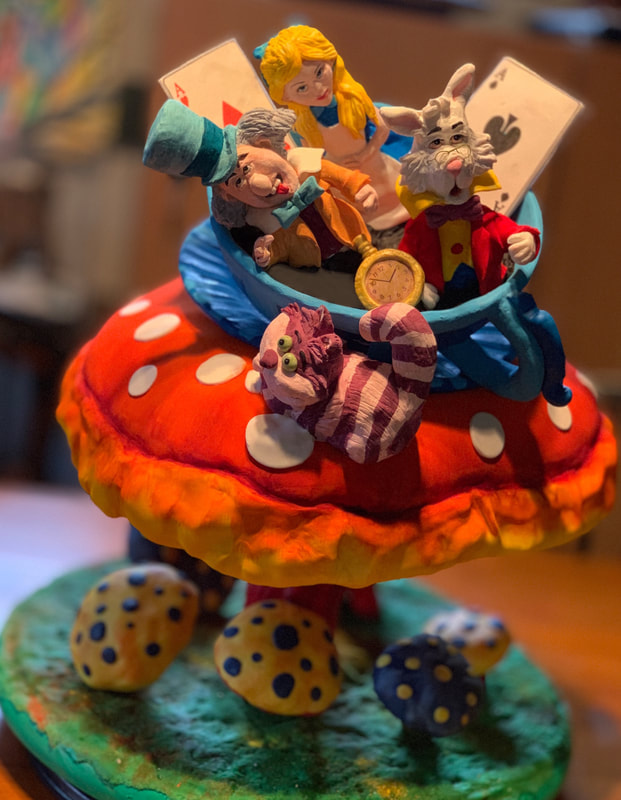 A whimsical sculpted cake depicts characters from Alice in Wonderland, including the Mad Hatter, the White Rabbit, and Alice, riding in a blue boat atop a large red mushroom, with the Cheshire Cat lounging on the cap and smaller spotted mushrooms decorating the green base.