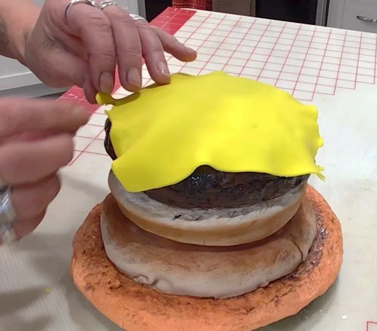 Hands place a thin yellow fondant cheese slice onto the burger patty.
