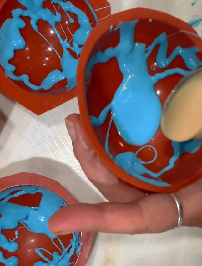 Hand pouring melted white chocolate into a silicone half-sphere mold.