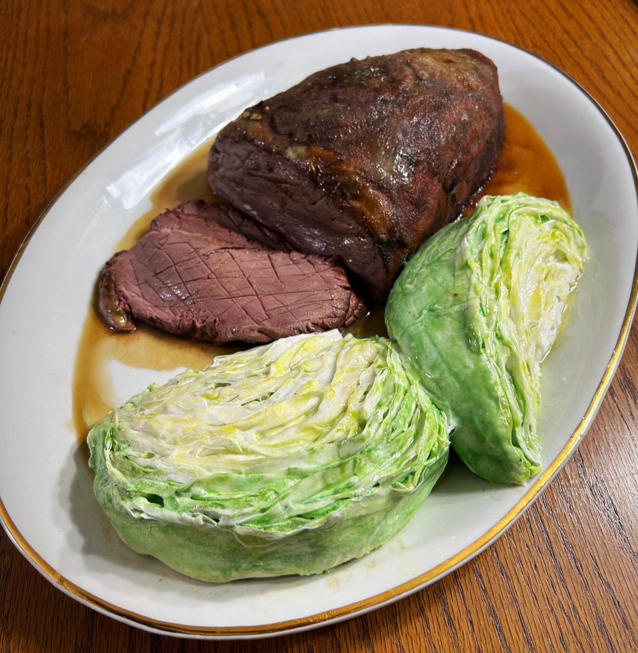 A hyper-realistic sculpted cake designed to look like a plated corned beef and cabbage dinner, with a dark roasted beef roast featuring a cross-hatched seared exterior and tender pink interior, two boiled cabbage halves with pale green and white layers, and brown gravy pooling around the base on an oval white platter with a gold rim.