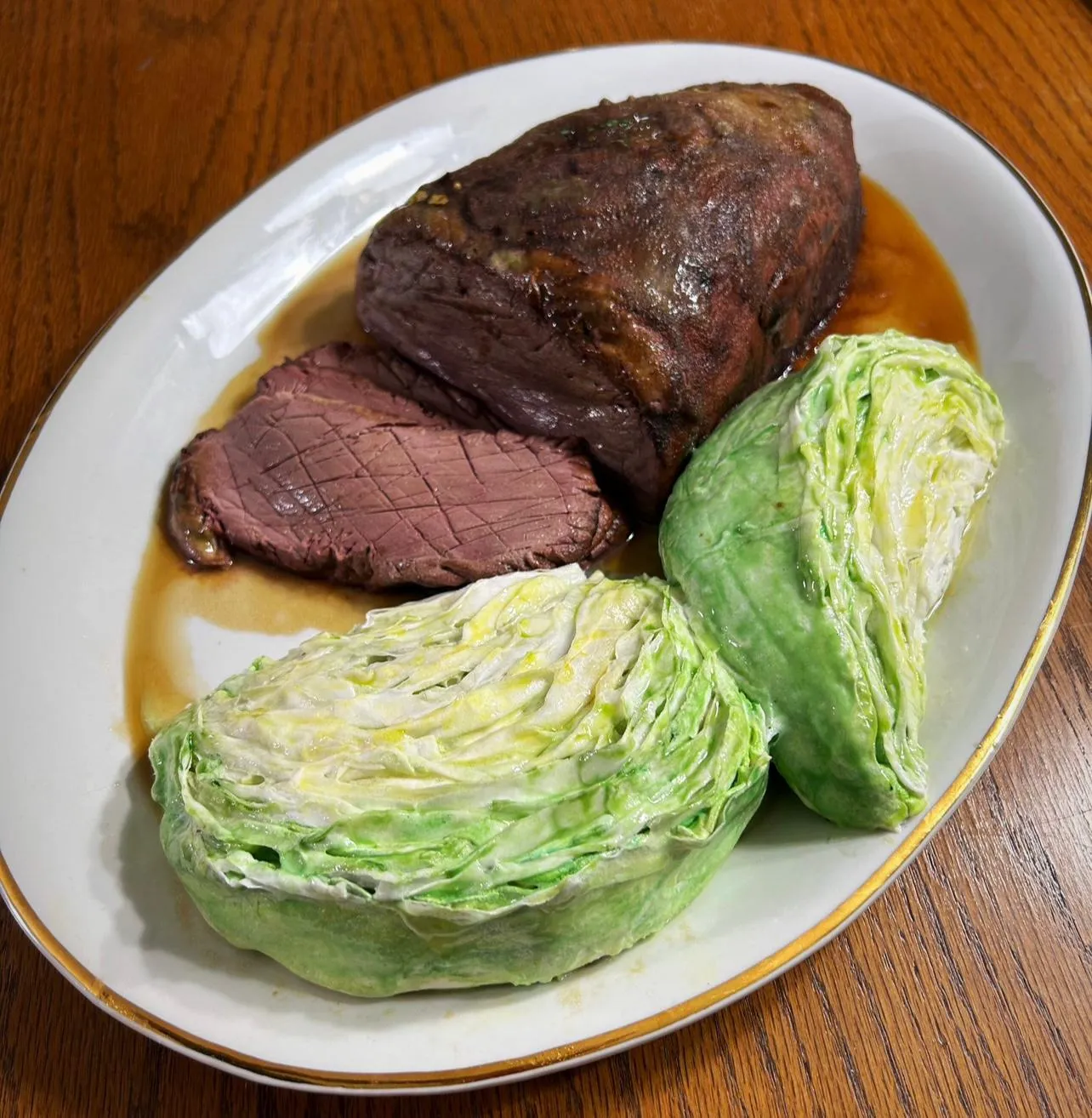 A hyper-realistic sculpted cake designed to look like a plated corned beef and cabbage dinner, with a dark roasted beef roast featuring a cross-hatched seared exterior and tender pink interior, two boiled cabbage halves with pale green and white layers, and brown gravy pooling around the base on an oval white platter with a gold rim.