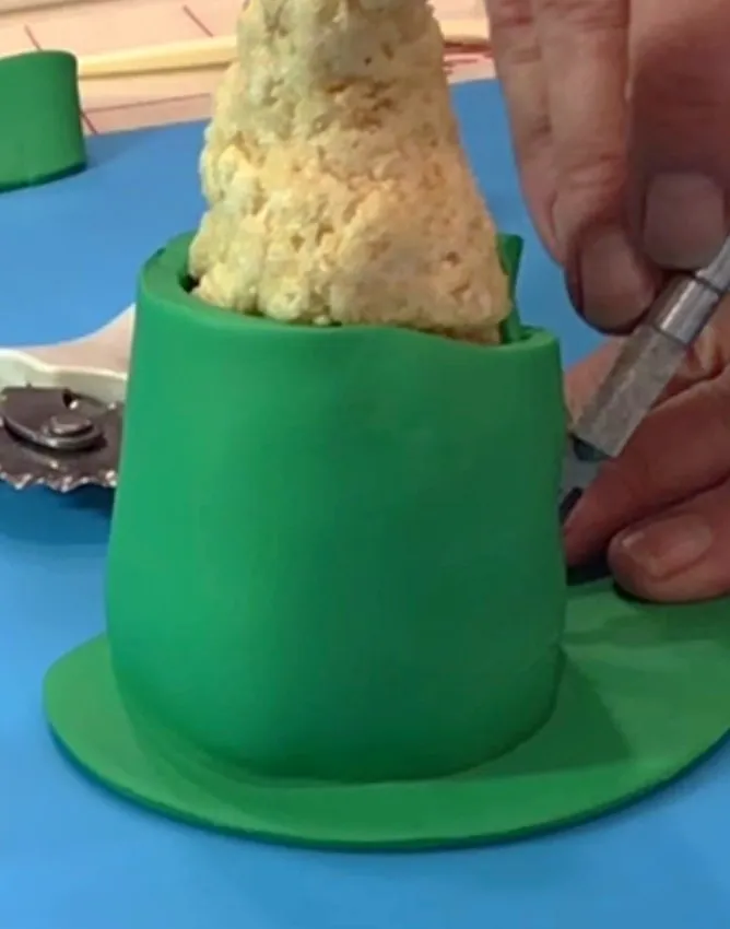 A hand trims green fondant covering the gnome body with a cake tool.
