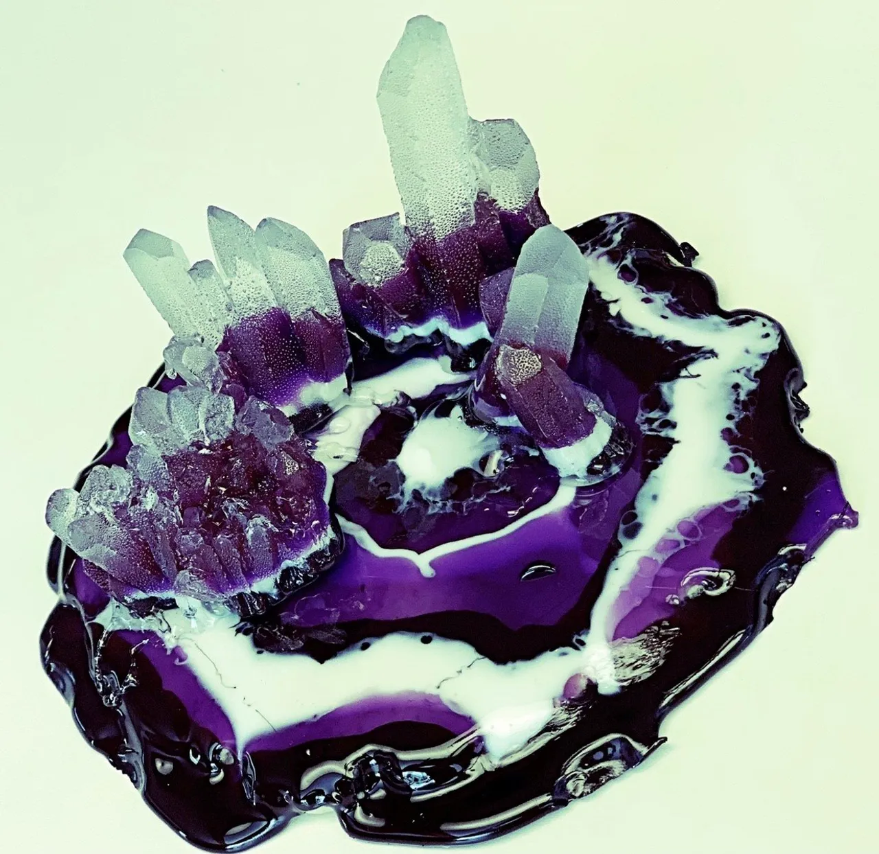 A sugar art piece featuring clear amethyst crystals with purple-dipped bases sits in a glossy pool of swirling purple and white spun sugar.