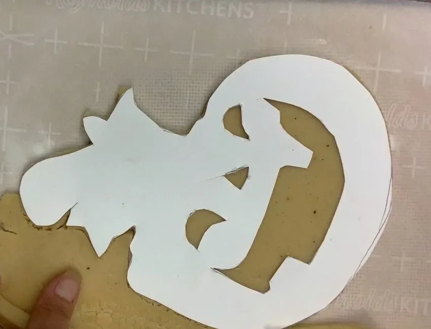A paper template and knife are used to cut Halloween cookie shapes from dough.