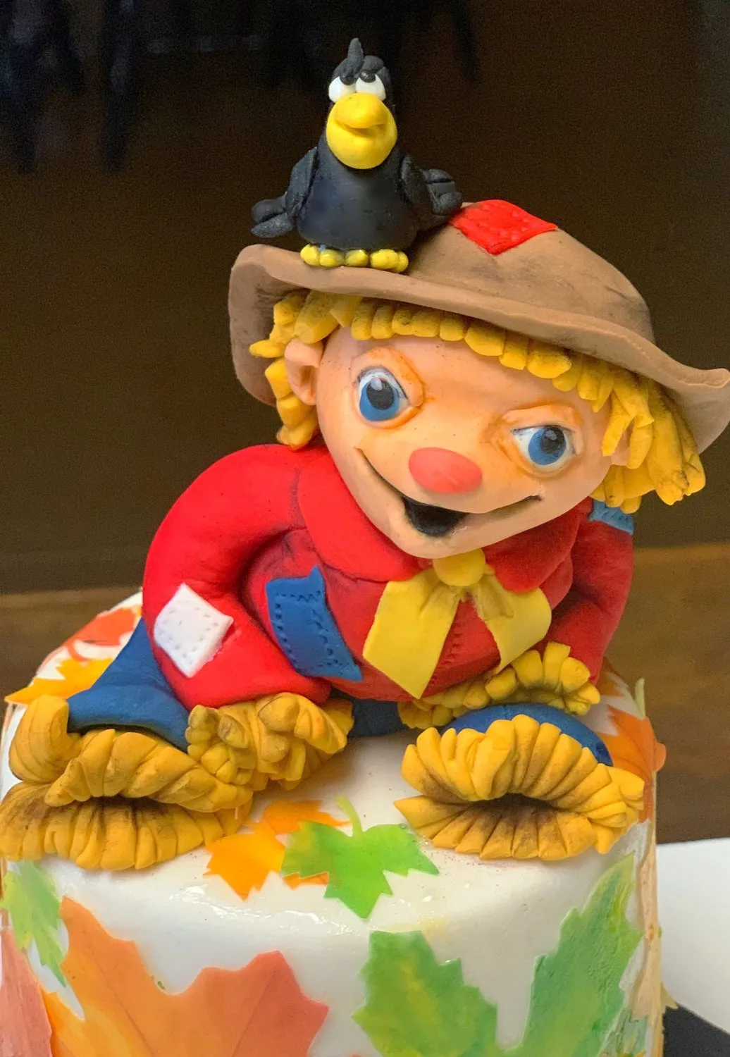 A detailed fondant cake topper features a smiling scarecrow with curly blonde hair, a red shirt, and a brown hat with a small black crow perched on it, set against a backdrop of colorful autumn leaves.