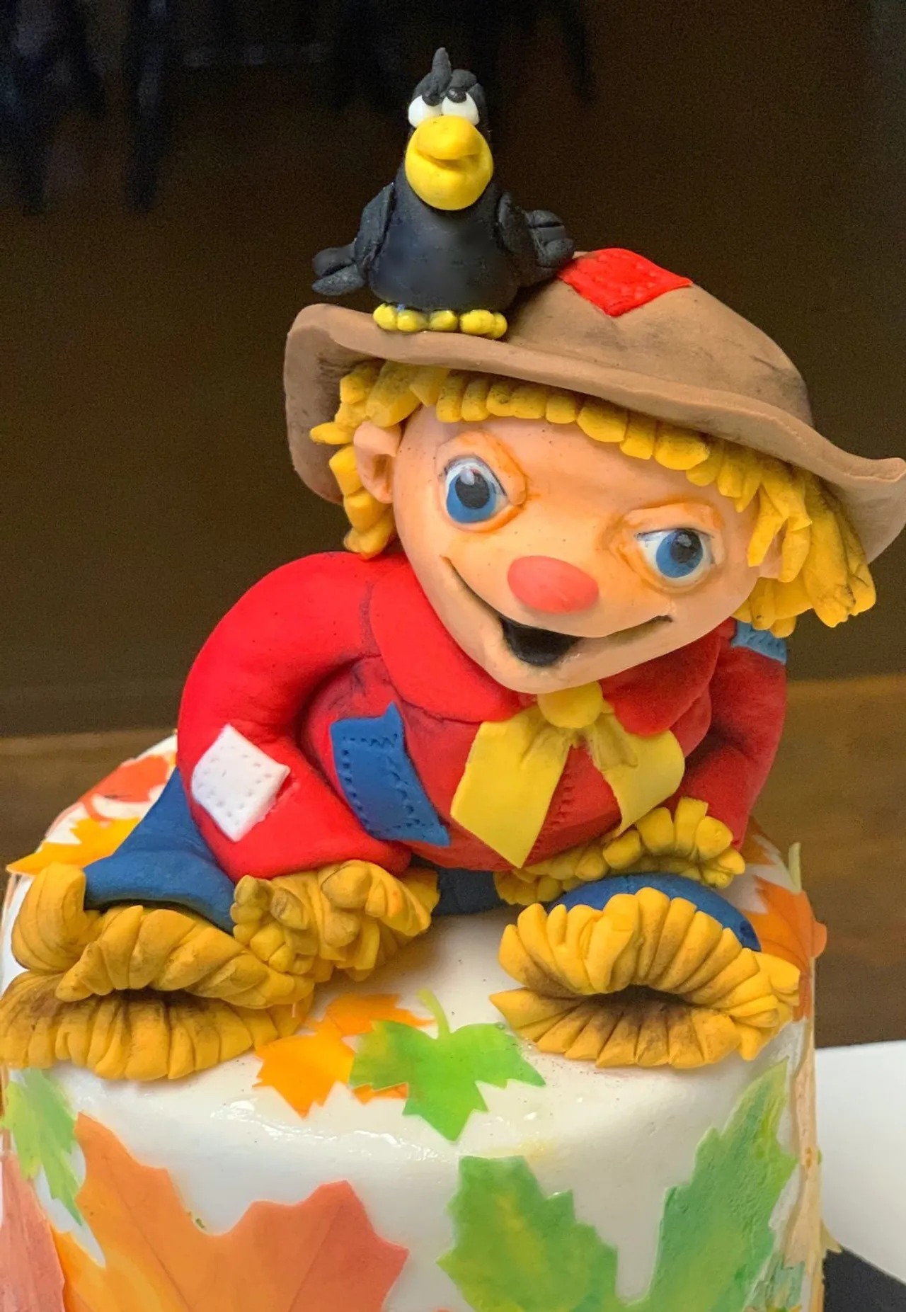 A detailed fondant cake topper features a smiling scarecrow with curly blonde hair, a red shirt, and a brown hat with a small black crow perched on it, set against a backdrop of colorful autumn leaves.