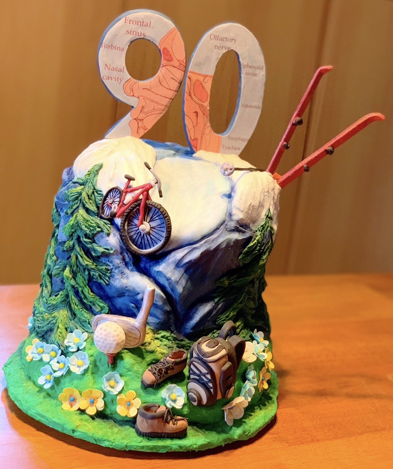 A sculpted cake tribute to Beetlejuice featuring the character seated atop mossy tombstone-adorned mound with spiky yellow hair, pale skin, dark eye makeup, black-and-white striped suit, large serpentine creature with striped neck and toothy head behind him, blue-skinned grinning face with wild orange hair on extending appendage, black-and-white striped base with "BEETLEJUICE" lettering and skull-and-crossbones emblem.
