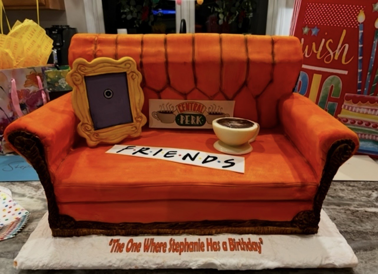 A sculpted cake replicating the iconic orange couch from Friends, with vibrant orange tufted backrest, framed picture, "Central Perk" sign, steaming coffee cup, text reading "The One Where Stephanie Has a Birthday."