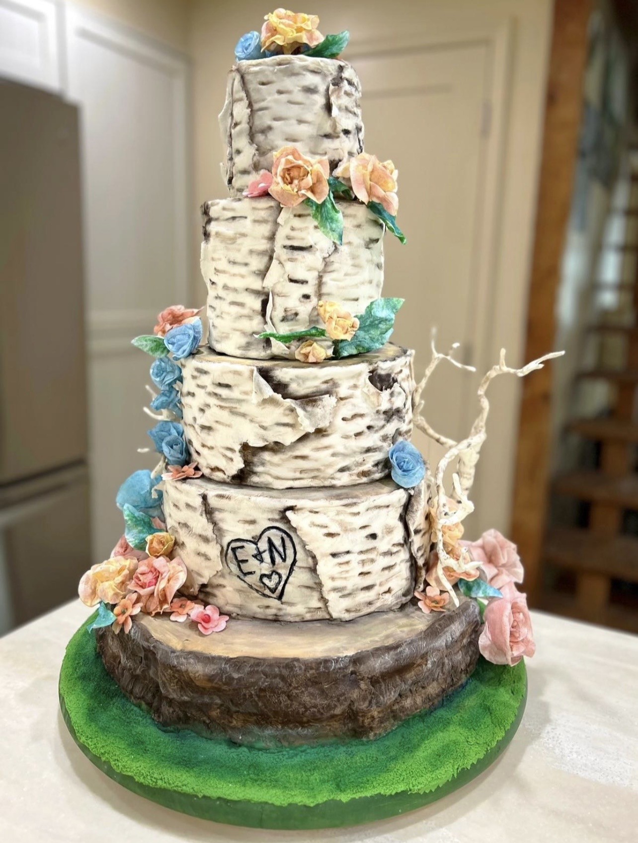 An intricately sculpted five-tiered wedding cake designed to resemble stacked birch tree logs resting on a mossy forest floor, each tier mimicking peeling papery bark with characteristic white surface and dark lenticels, decorated with delicate sugar flowers in blush pink, peach, sky blue, and pale yellow, with a hand-drawn heart containing the initials "E & N" on the second-lowest tier.