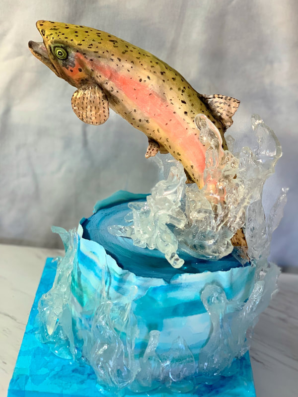 A hyper-realistic cake features a detailed trout leaping from a blue, wave-like base, surrounded by translucent, crystal-like sugar splashes.