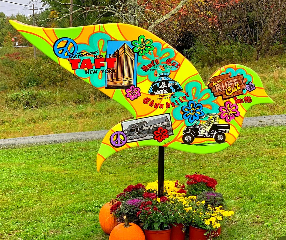 A large, colorful, bird-shaped sign with a psychedelic 1960s design stands in a grassy area, featuring illustrations and text for local businesses like "The Famous Hotel Taft New York" and "Annie's Ruff Cut," surrounded by potted mums and pumpkins.