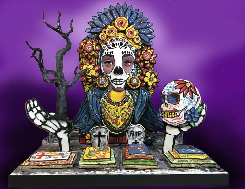 A sculpted cake figure of a female figure with Day of the Dead skull makeup and a vibrant floral headdress holds a decorated skull in one hand and a bare branch in the other, standing behind an altar with sugar skulls, tombstones marked "RIP," and colorful offerings.