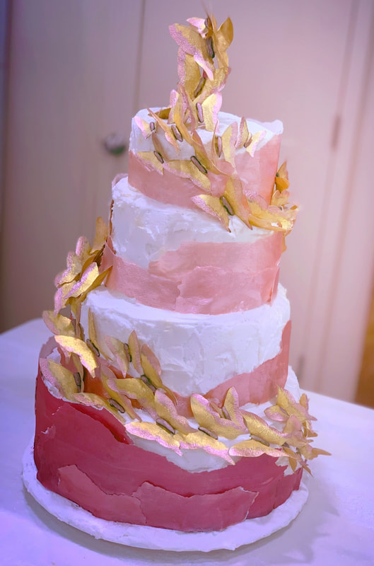 A four-tiered wedding cake features textured white frosting with sweeping bands of pink and red icing, adorned with cascading gold leaf decorations that spiral from the top tier down to the base.