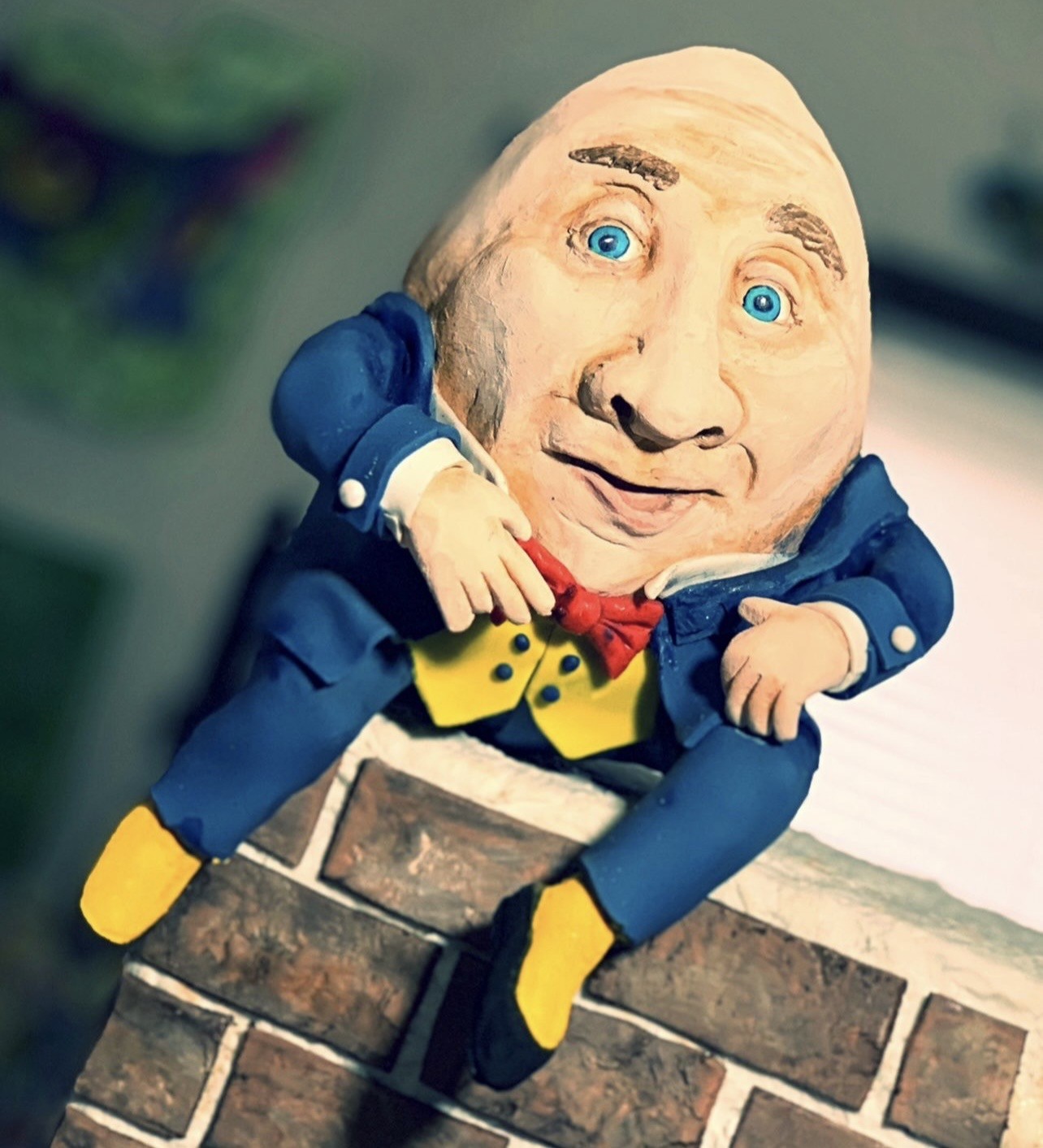 A sculpted cake figurine of a man with an oversized expressive head, wide bright blue eyes, and a knowing smirk, wearing a royal blue tailcoat with white cuffs, vibrant yellow waistcoat with dark buttons, bold red bow tie, seated on a brick ledge.