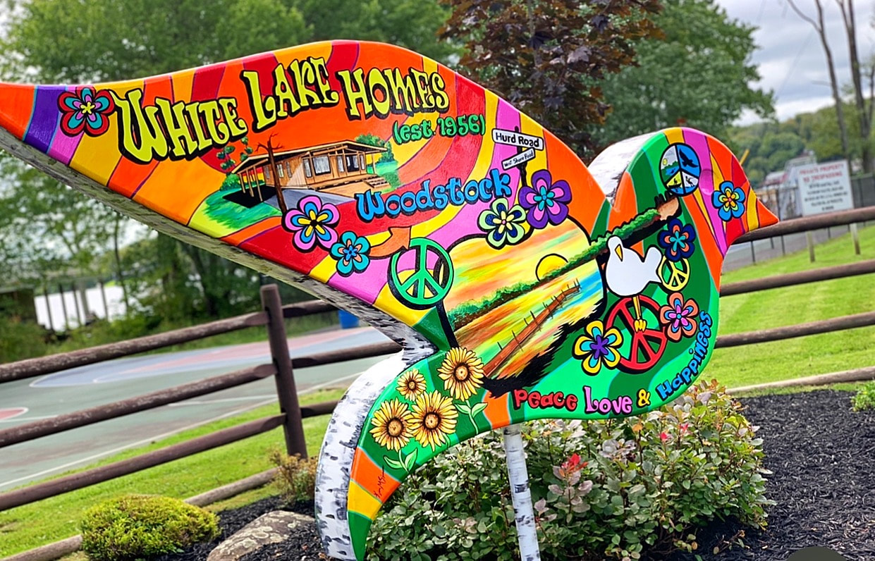 A colorful, psychedelic bird sculpture painted with peace signs, flowers, and sunflowers features the text "White Lake Homes (est. 1956) Woodstock" and "Peace Love & Happiness" while standing in a landscaped garden bed.