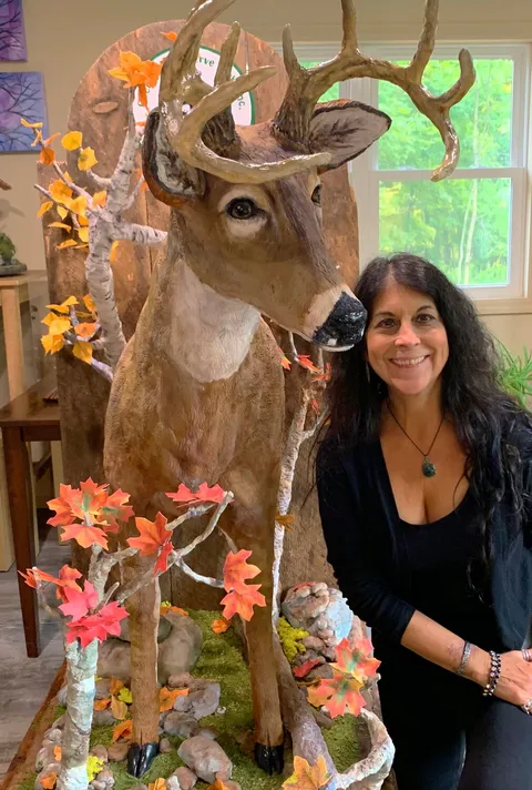 Kim Simons standing beside a life-sized deer cake sculpture in an autumnal scene.
