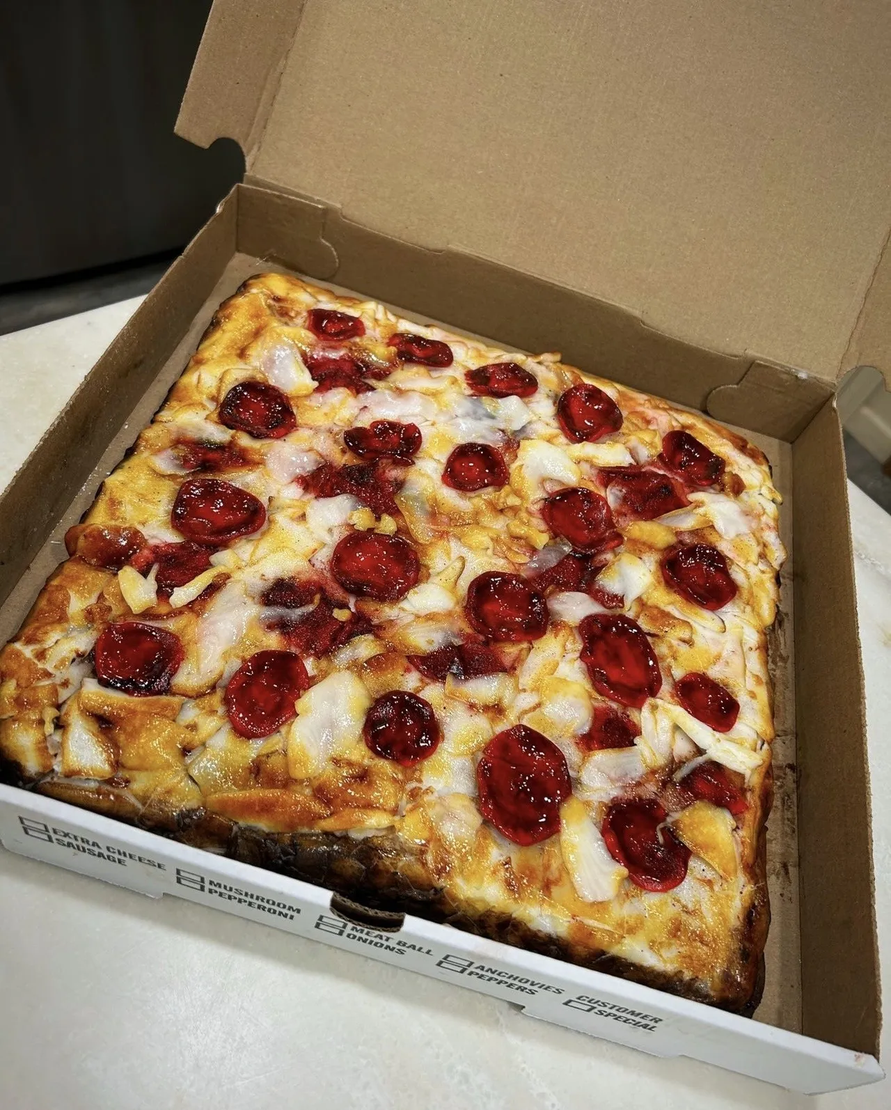A sculpted cake designed as a freshly baked square pepperoni pizza inside an open cardboard box, with melted cheese bubbles, glossy curled pepperoni slices glistening with rendered fat, white onion pieces peeking through, and a golden-brown crust.