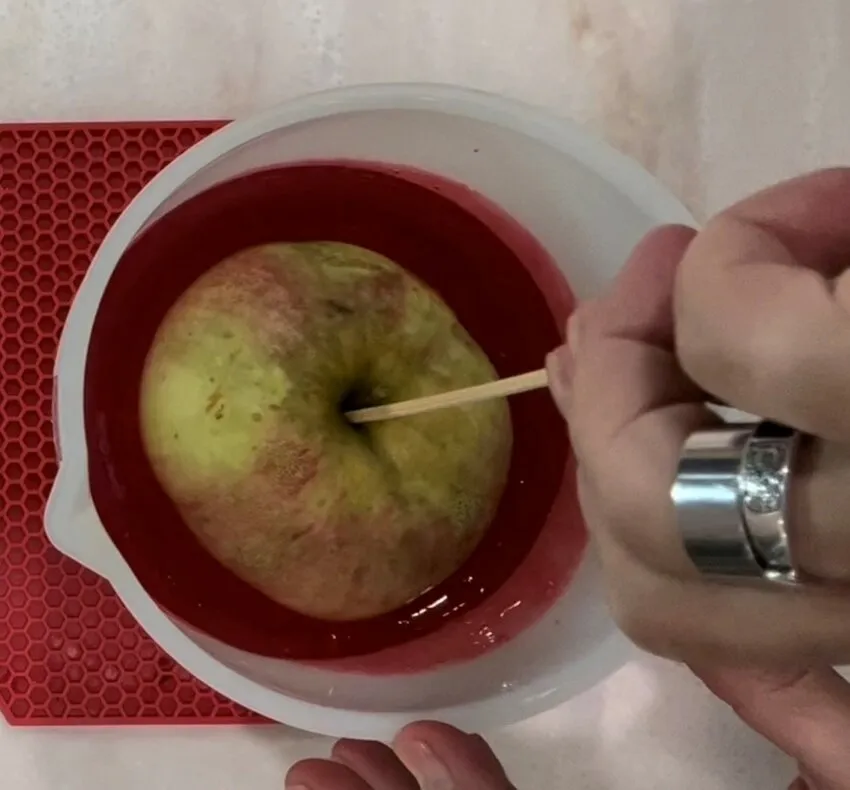 A hand dips a skewered apple into melted red candy coating.