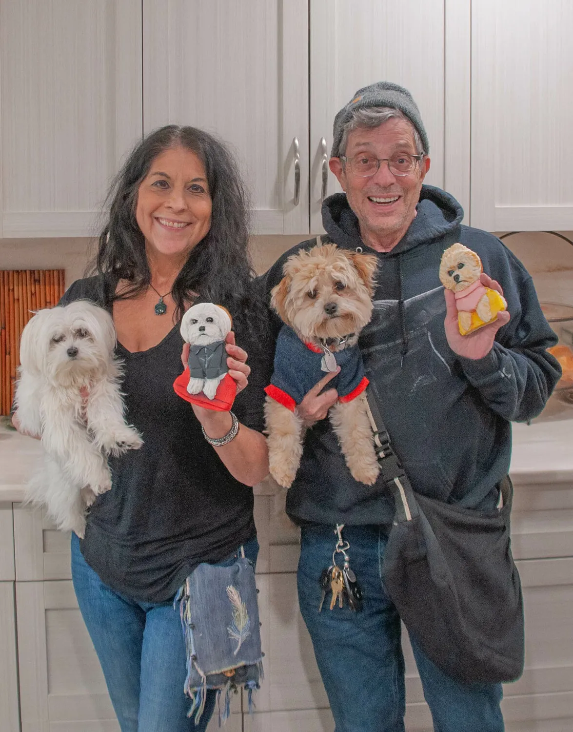 Two people each holding a small white dog, with decorated pet-shaped sugar cookies displayed in front