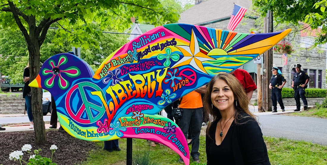 A smiling woman stands next to a large, colorful bird-shaped sign painted in a psychedelic style, featuring peace symbols and the word "LIBERTY" surrounded by names of local businesses like "Sullivan's" and "Liberty Theater."