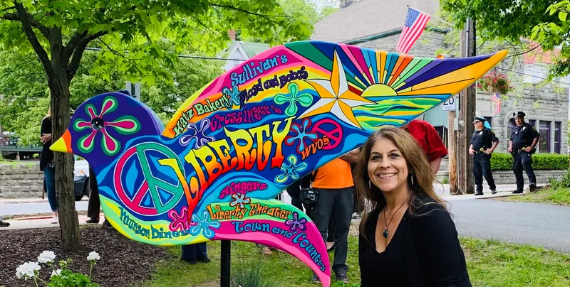 A smiling woman stands next to a large, colorful bird-shaped sign painted in a psychedelic style, featuring peace symbols and the word "LIBERTY" surrounded by names of local businesses like "Sullivan's" and "Liberty Theater."
