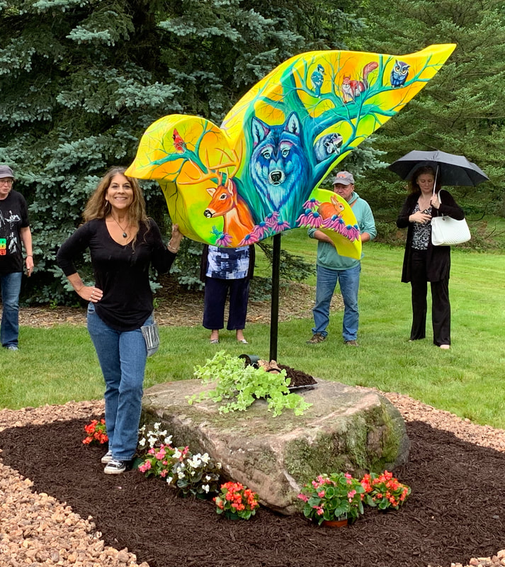 A smiling woman in a black shirt and jeans stands next to a colorful, bird-shaped sign for the "Reel Harmony Songwriter Festival" that features musical instruments and the words "Peace Love and Music."