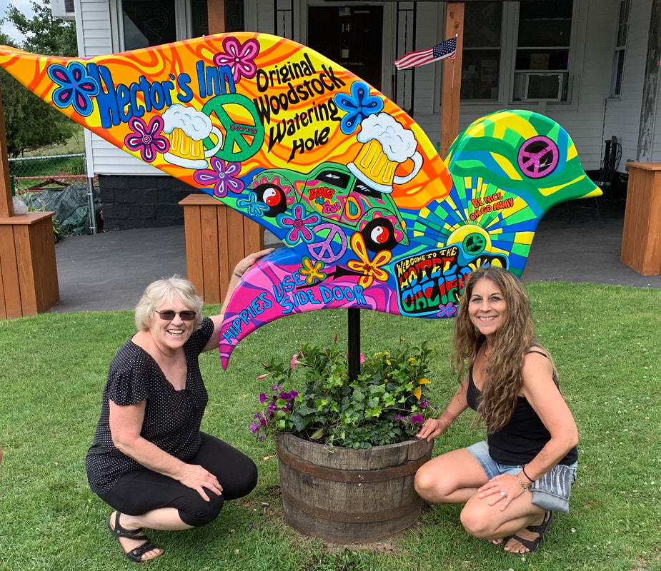 Two women crouch on a lawn beside a colorful, psychedelic bird-shaped sign for "Hector's Inn Original Woodstock Watering Hole" that features peace signs, flowers, and beer mugs.
