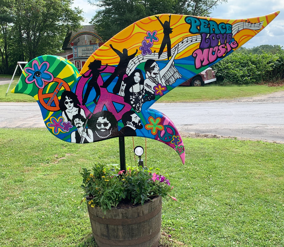 A colorful, psychedelic-style wooden cutout shaped like a bird or leaf, featuring silhouettes of musicians, peace signs, flowers, and the words "PEACE LOVE MUSIC" in vibrant lettering, mounted on a post above a barrel planter filled with blooming flowers.