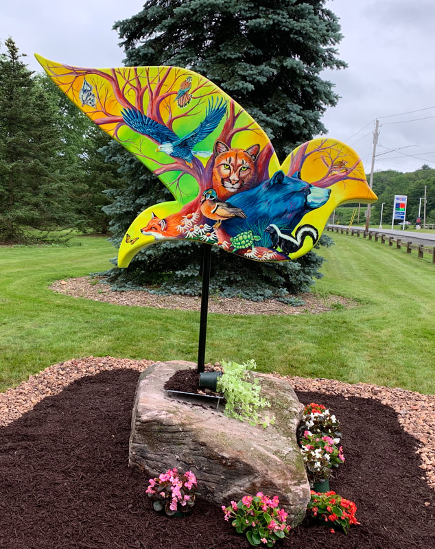 A vibrant, bird-shaped wooden sculpture painted with a colorful collage of wildlife—including a cougar, bear, fox, and various birds—stands mounted on a black pole atop a large rock, surrounded by mulch and small potted flowers in a grassy roadside setting.