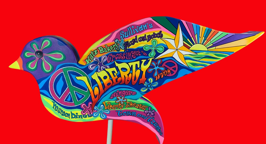 A smiling woman stands beside a large, colorful bird-shaped sign for the "Lenape Wurtsboro D & H Canal," which features vibrant rainbow stripes, painted landscapes, and wildlife symbols.