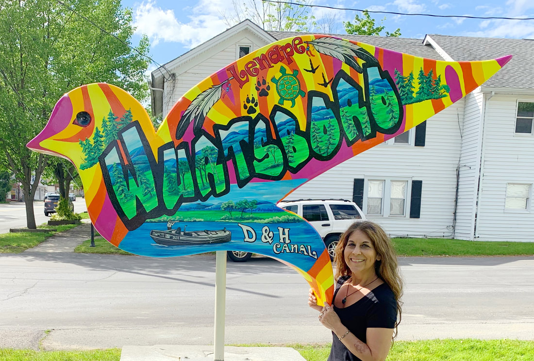 A colorful bird-shaped wooden sign features a vibrant forest scene with a blue wolf, deer, owl, raccoon, squirrel, and various birds nestled among trees and purple flowers.
