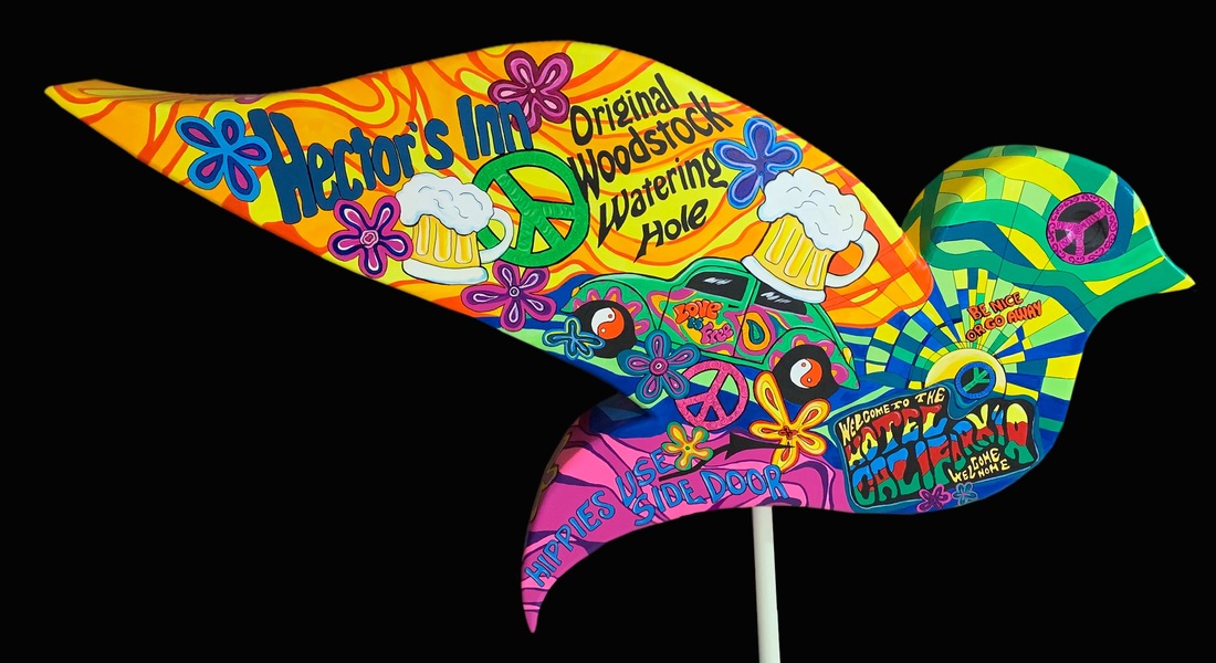 A large, vibrantly painted bird sculpture stands in a wooden planter box, featuring colorful peace signs, handprints, and text reading "Walmart Mountain," "Soap Box Derby," and "Liberty Museum & Arts Center."