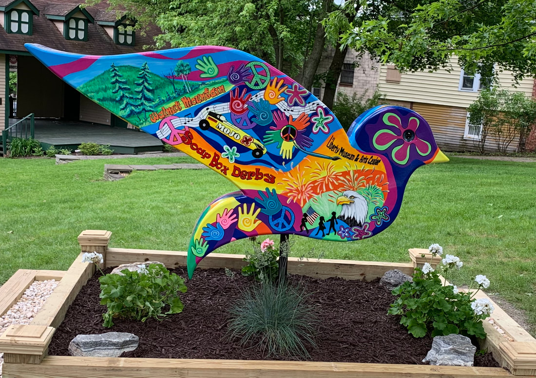 A woman in a black shirt and jeans stands smiling beside a large, colorful butterfly sculpture featuring a wolf, deer, and birds, mounted on a rock surrounded by flowers in a garden setting with other people in the background.