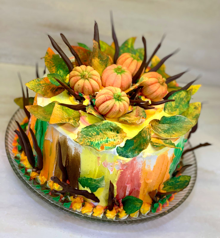 A round cake decorated with colorful autumn-themed frosting, featuring small orange pumpkins, green and yellow leaves, and dark chocolate spikes arranged on top and around the sides.
