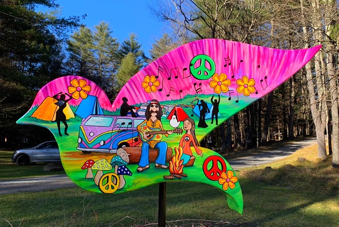 A vibrant, psychedelic mural shaped like a mustache depicts a 1960s hippie scene featuring a couple playing guitar by a campfire, a colorful VW bus, tents, peace signs, and flowers.