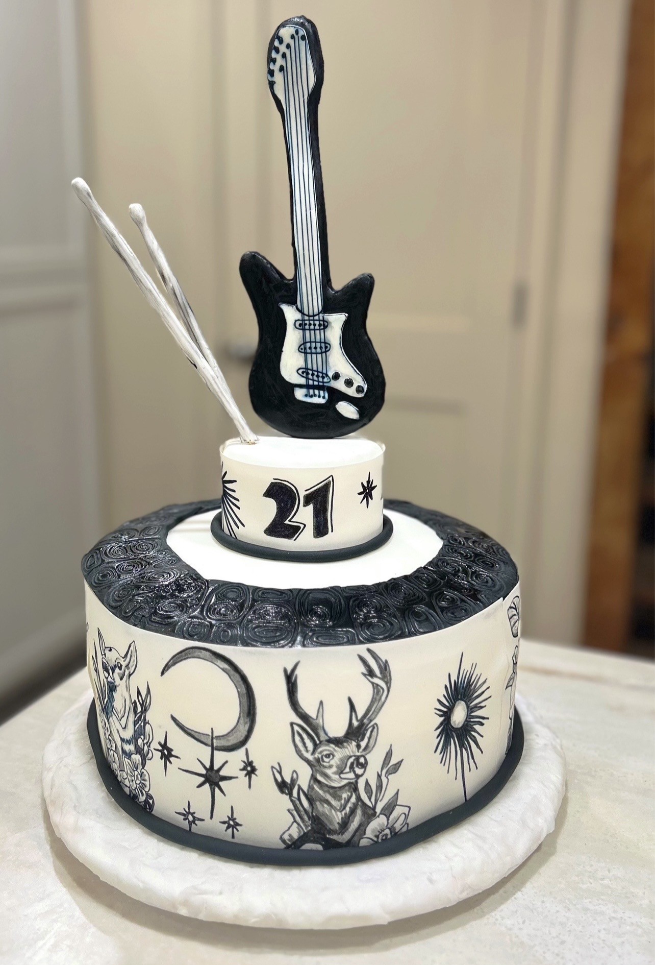 A two-tiered rock-and-roll themed cake with monochromatic black and white design — lower tier in smooth white fondant with black ink-style illustrations of crescent moon, sunburst, stars, two deer making peace signs, floral motifs, textured black band with swirling patterns, upper tier displaying bold number "21" flanked by starbursts, topped by meticulously crafted black electric guitar with white pickguard and two silver-painted drumsticks.