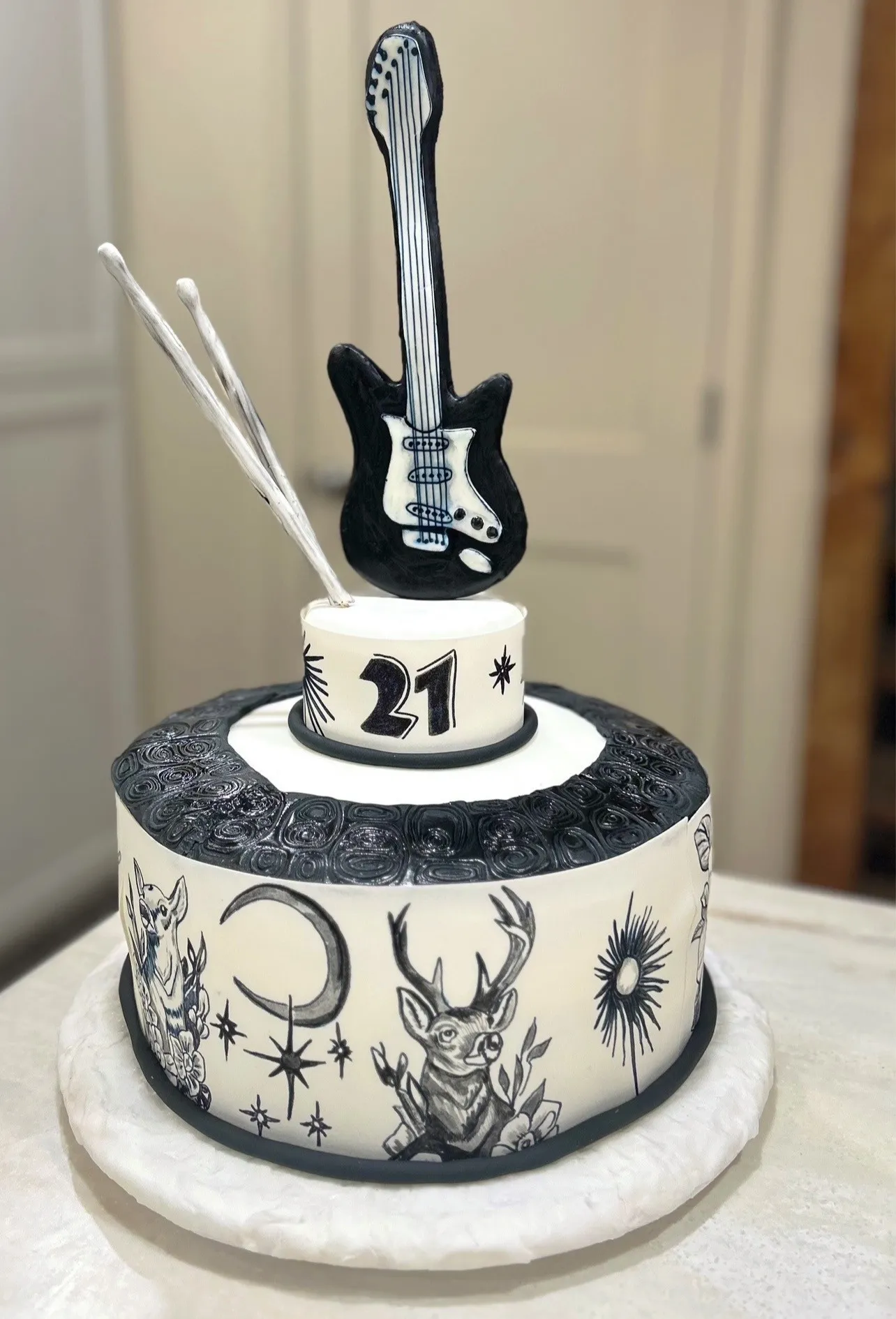 A two-tiered rock-and-roll themed cake with monochromatic black and white design \u2014 lower tier in smooth white fondant with black ink-style illustrations of crescent moon, sunburst, stars, two deer making peace signs, floral motifs, textured black band with swirling patterns, upper tier displaying bold number "21" flanked by starbursts, topped by meticulously crafted black electric guitar with white pickguard and two silver-painted drumsticks.