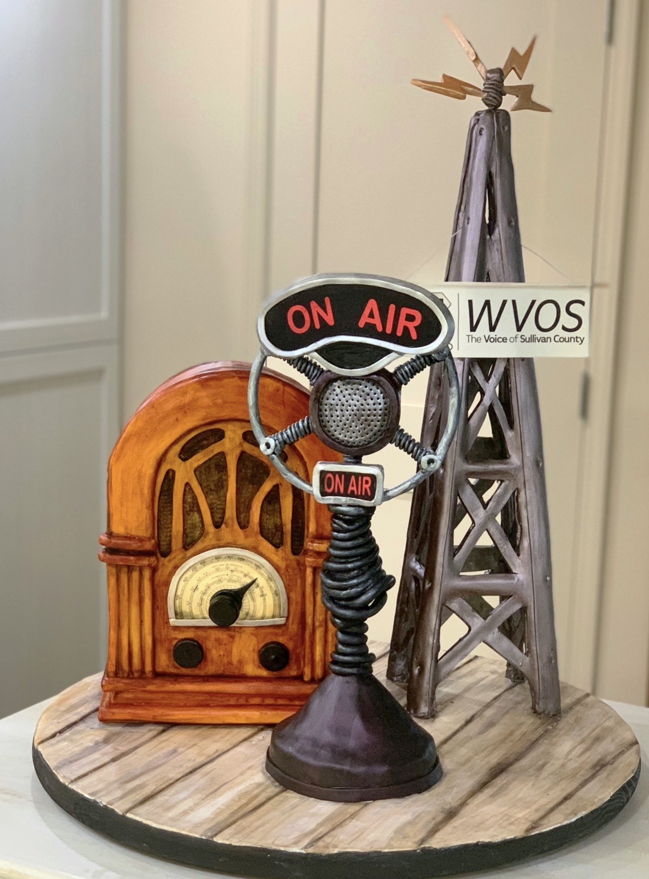 A sculpted cake diorama of a 1940s noir detective's desk featuring faux leather-bound book base, cream-colored fedora with black band, crumpled newspaper titled "DAILY NEWS" with headline "Breaking News: Mike Retires," metallic retro-style microphone, binoculars, on warm-toned wooden table.
