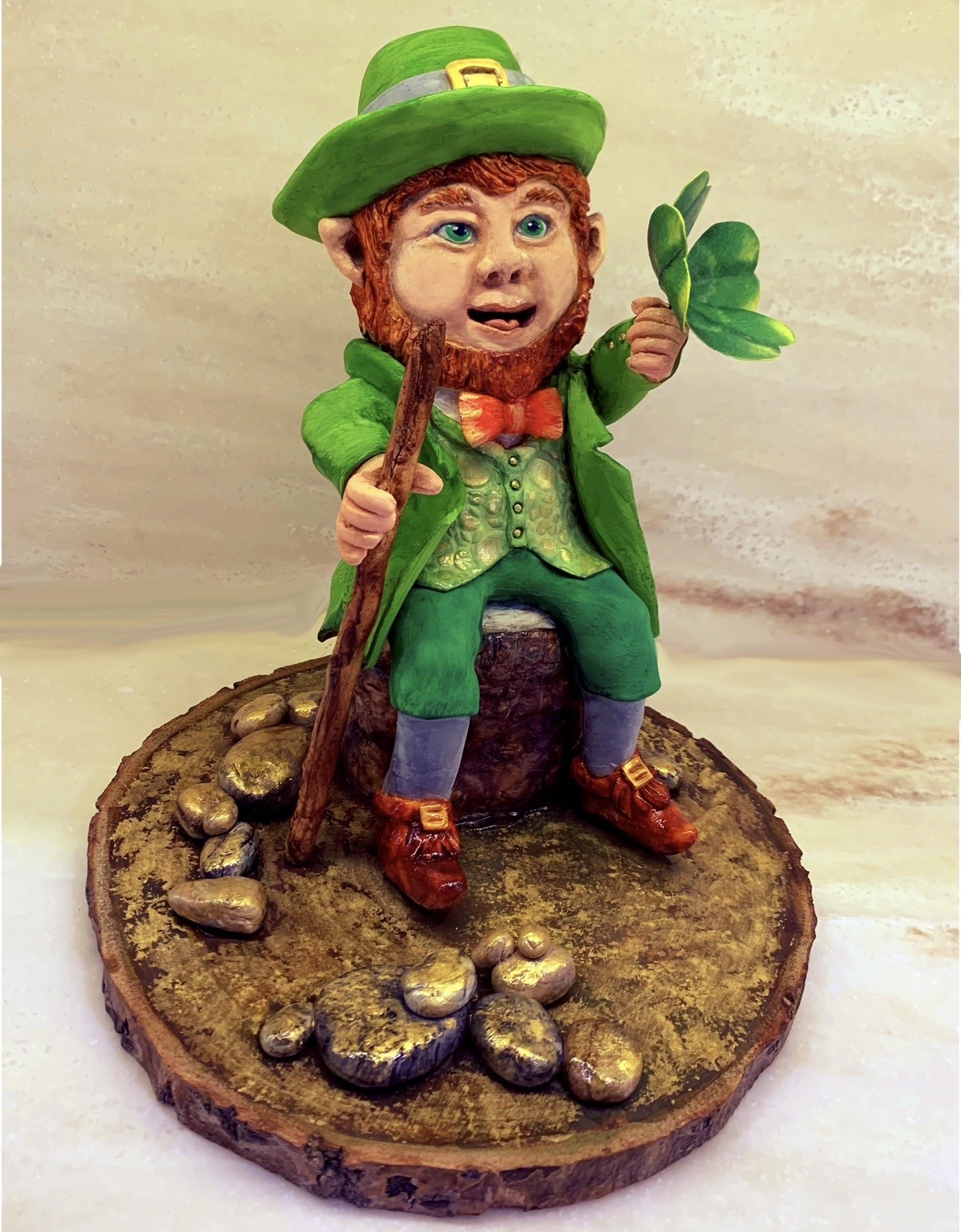 A St. Patrick's Day sculpted cake figurine of a leprechaun seated on a rustic wooden slice base, wearing a vibrant green suit with textured vest, wide-brimmed green hat with silver band and gold buckle, reddish-brown beard, cheerful animated face, holding a wooden staff and bright green four-leaf clover, red bow tie, brown shoes with gold buckles, scattered gold-painted coins.