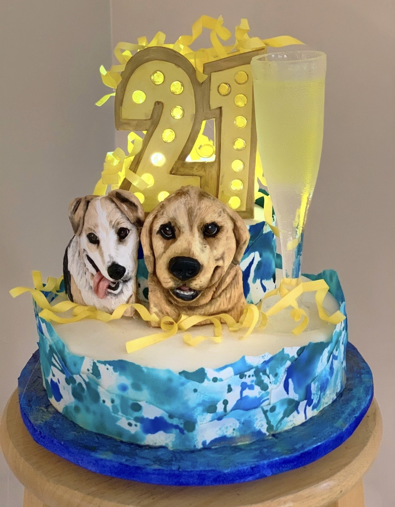 A 21st birthday cake with aquatic theme featuring dynamic blue watercolor-style frosting transitioning from deep navy to turquoise, topped by two realistic edible fondant portraits of dogs — a white and tan terrier mix and a golden Labrador — with a large illuminated marquee "21" in gold adorned with glowing yellow bulbs, curly ribbon streamers, and a translucent fondant champagne flute.