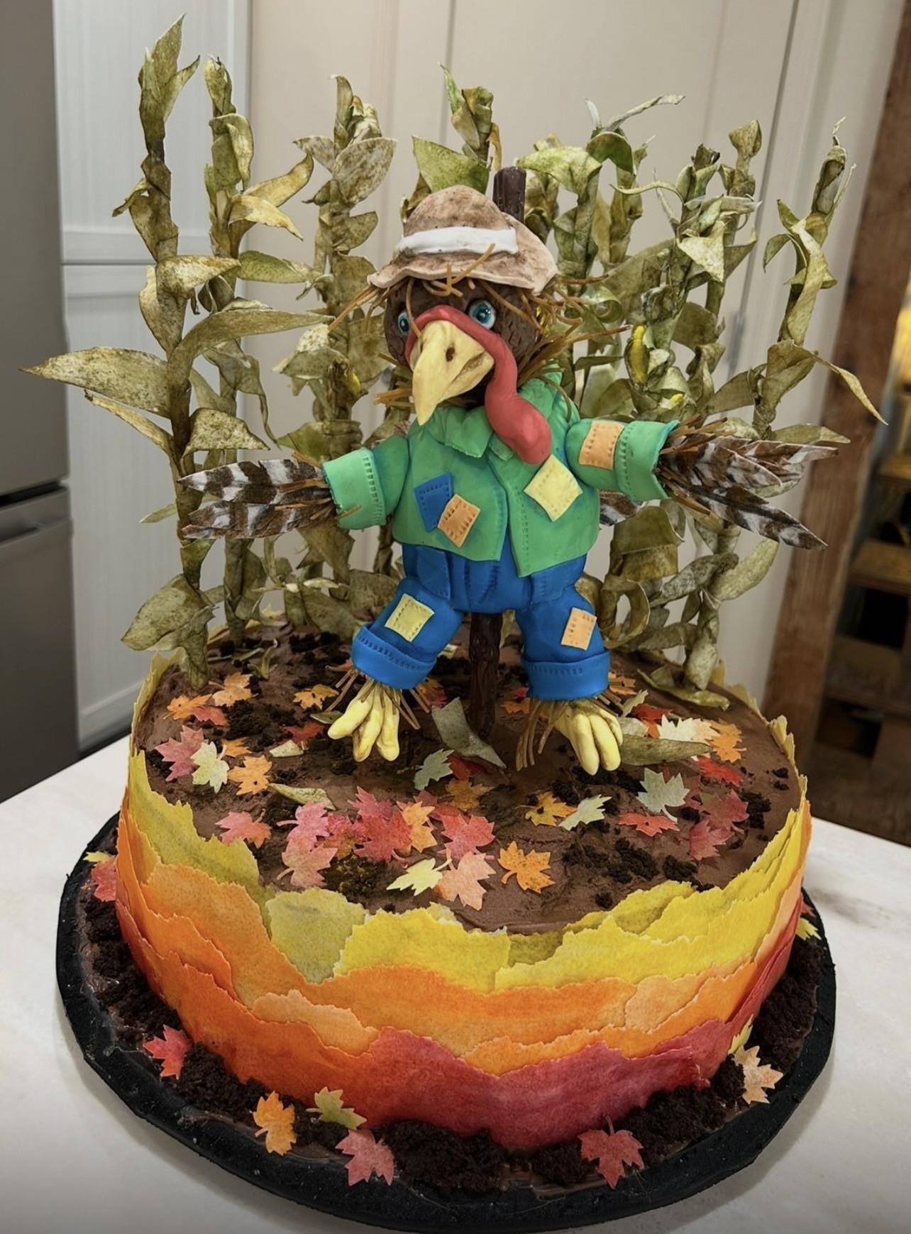 A Thanksgiving sculpted cake depicting a scarecrow-like turkey standing in a field of corn — the turkey with brown body, blue eyes, red wattle, yellow beak, green jacket with patchwork details, blue pants, straw hat with white band, wings revealing feathered patterns, surrounded by tall dried corn stalks, chocolate-frosted cake base with translucent fondant autumn border in red, orange, yellow, and green.