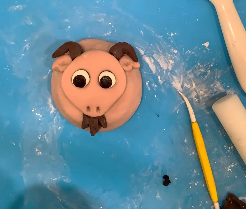 A pink fondant sheep head with brown horns, beard, and facial features.