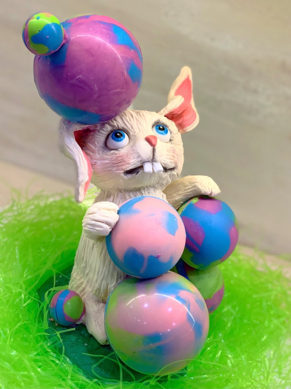 A whimsical sculpted cake of a white rabbit with blue eyes and buck teeth balances a stack of pastel-colored, swirled Easter eggs on its head and in its paws while sitting in a bed of bright green Easter grass.