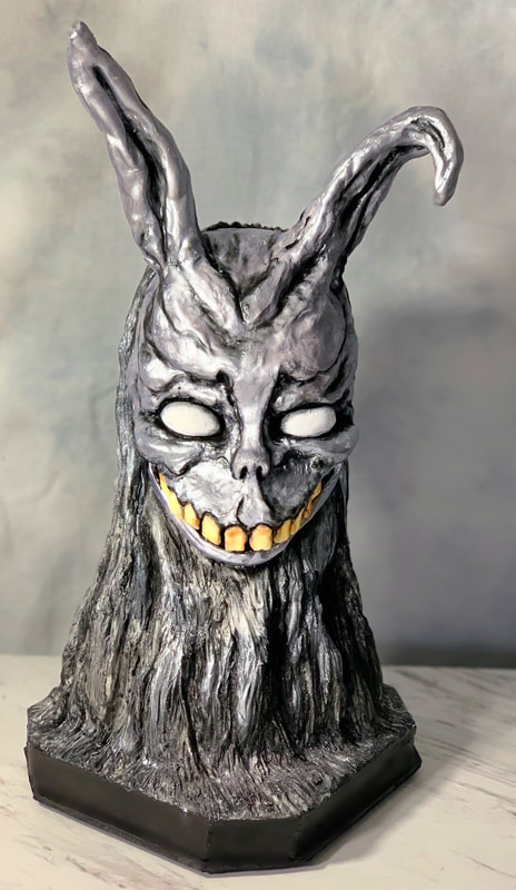 A sculpted cake bust of a silver-colored rabbit-like creature with long ears, glowing white eyes, and a wide, toothy grin, mounted on a black hexagonal base.