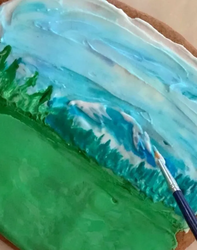 Hand detailing the mountain and field scene on the cookie with a brush.