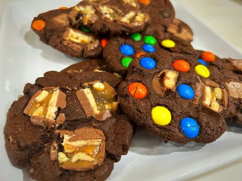Several dark chocolate cookies on a white plate, topped with colorful M&Ms and candy bar pieces