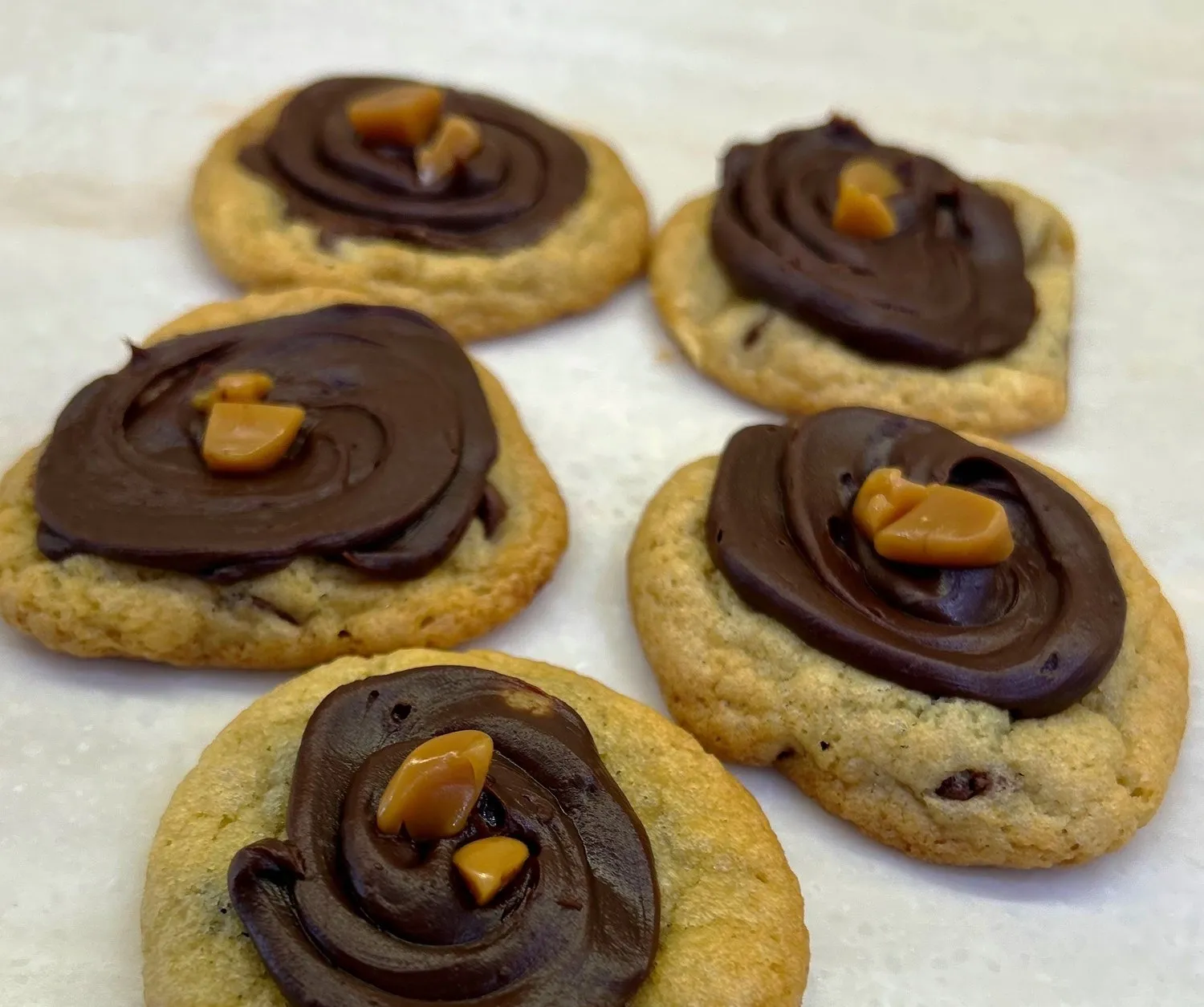 Round chocolate chip cookies topped with swirled chocolate frosting and caramel pieces on a white surface