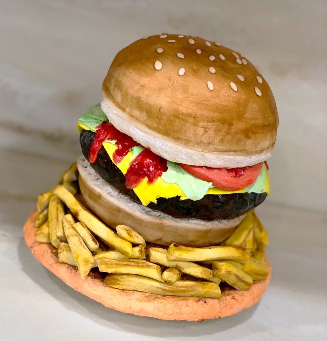 A hyper-realistic sculpted cake of a classic cheeseburger and fries — thick dark brown beef patty with bright yellow melted cheese and red ketchup, layered between pale green lettuce and red tomato slice, topped with a golden-brown sesame seed bun, all on a platter piled with golden-brown french fries.