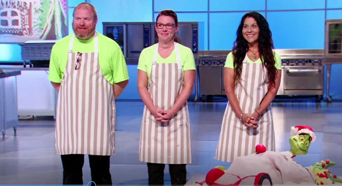 Three contestants in matching lime green shirts and striped aprons standing next to a Grinch-themed cake on Cake Wars Christmas.